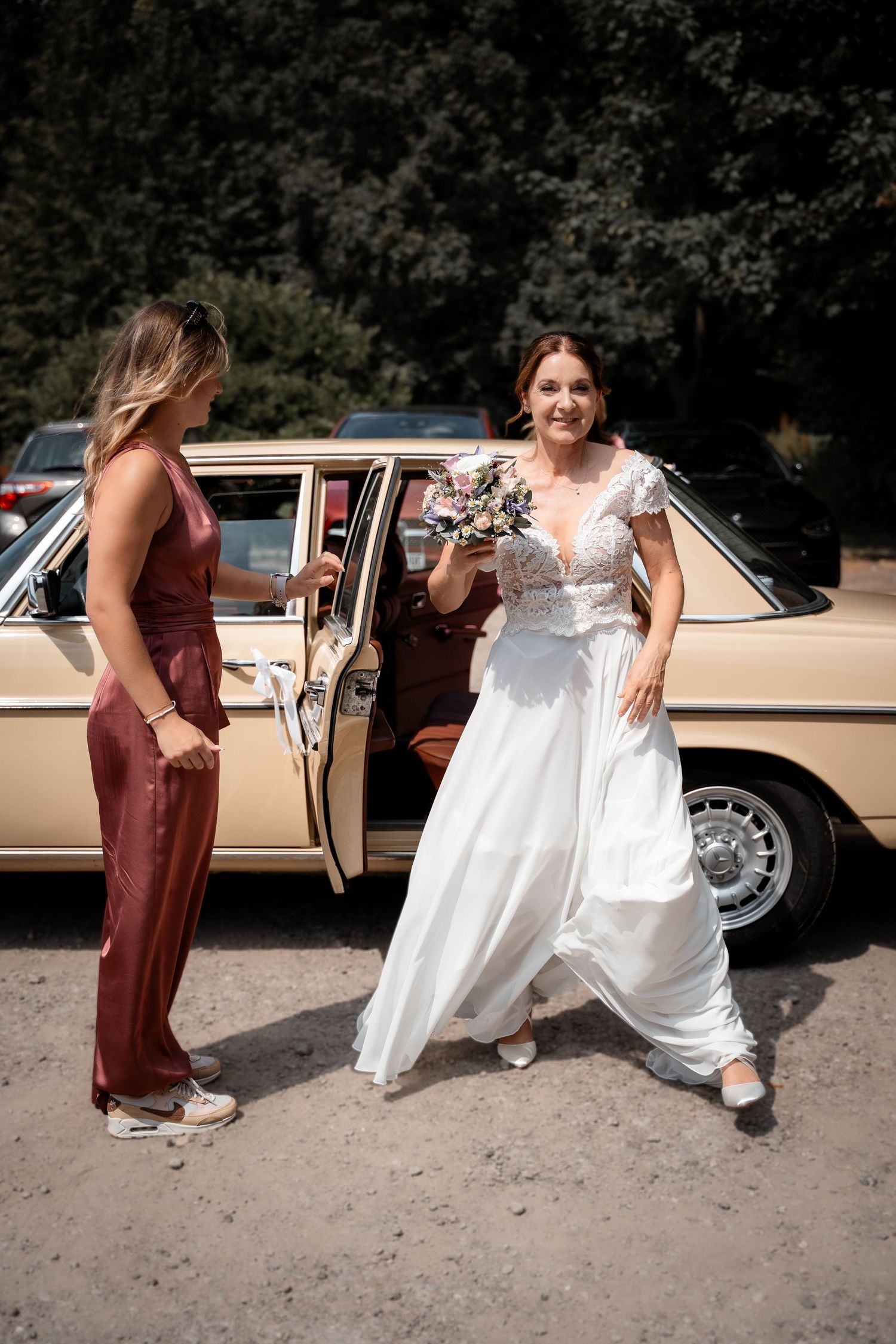 Bride in white lace gown stepping out of vintage beige car, holding bouquet, assisted by woman in red satin jumpsuit.