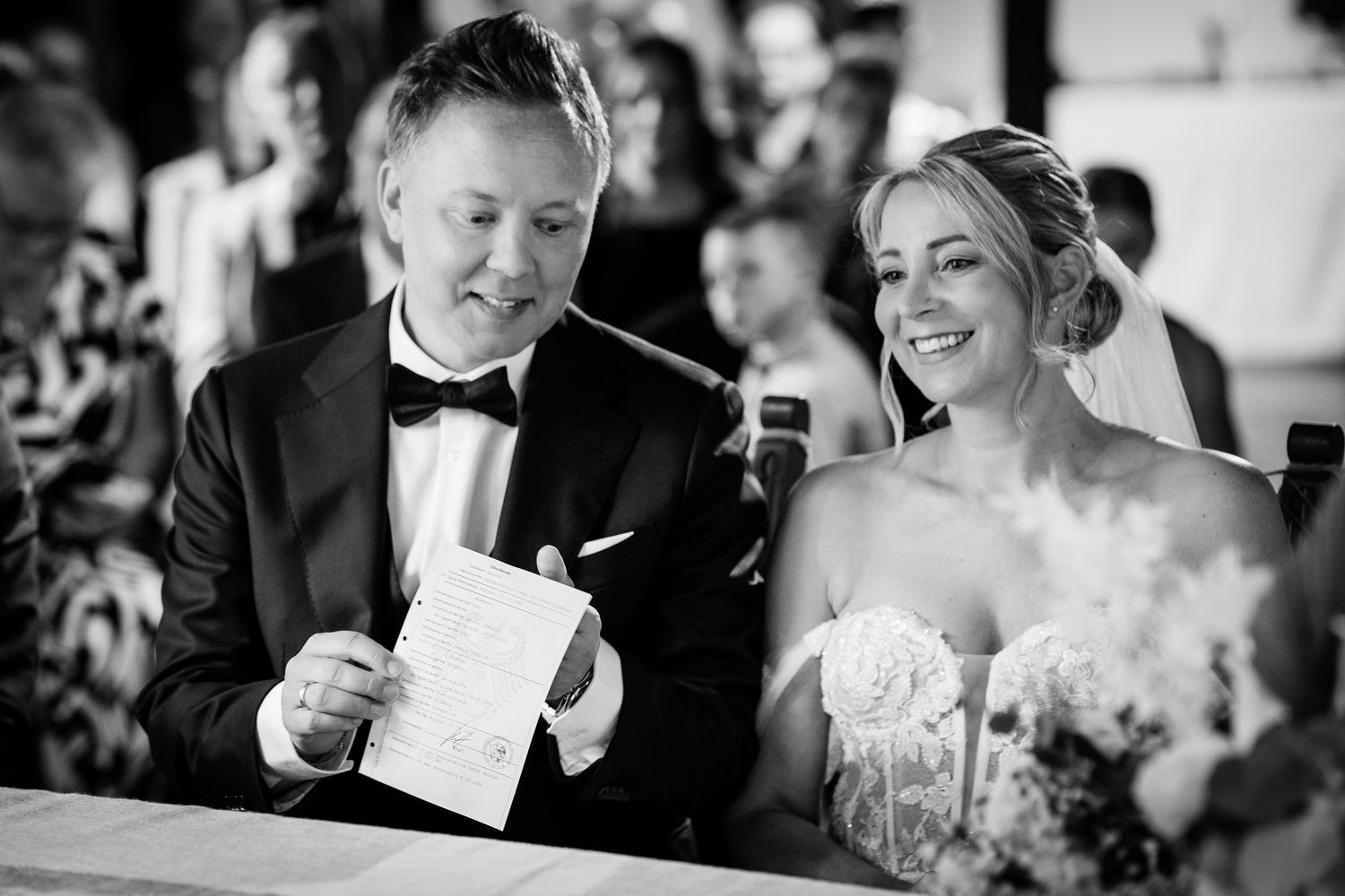 Bride laughing with groom holding paper during wedding ceremony in black and white.