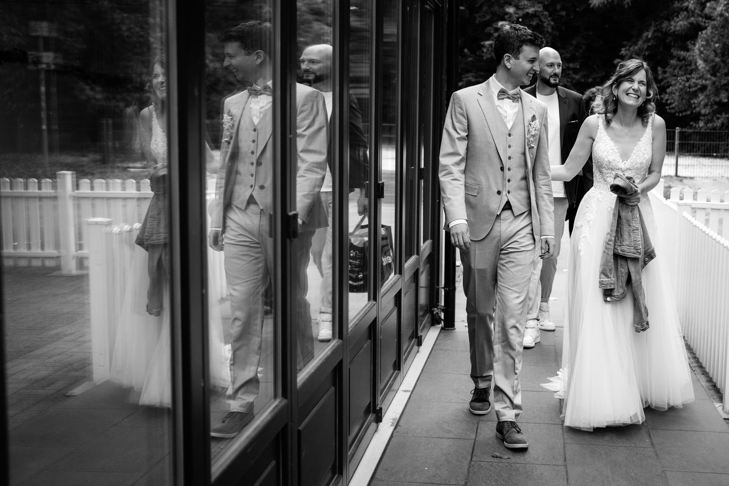 Black and white photo of bride and groom walking hand in hand past glass windows.