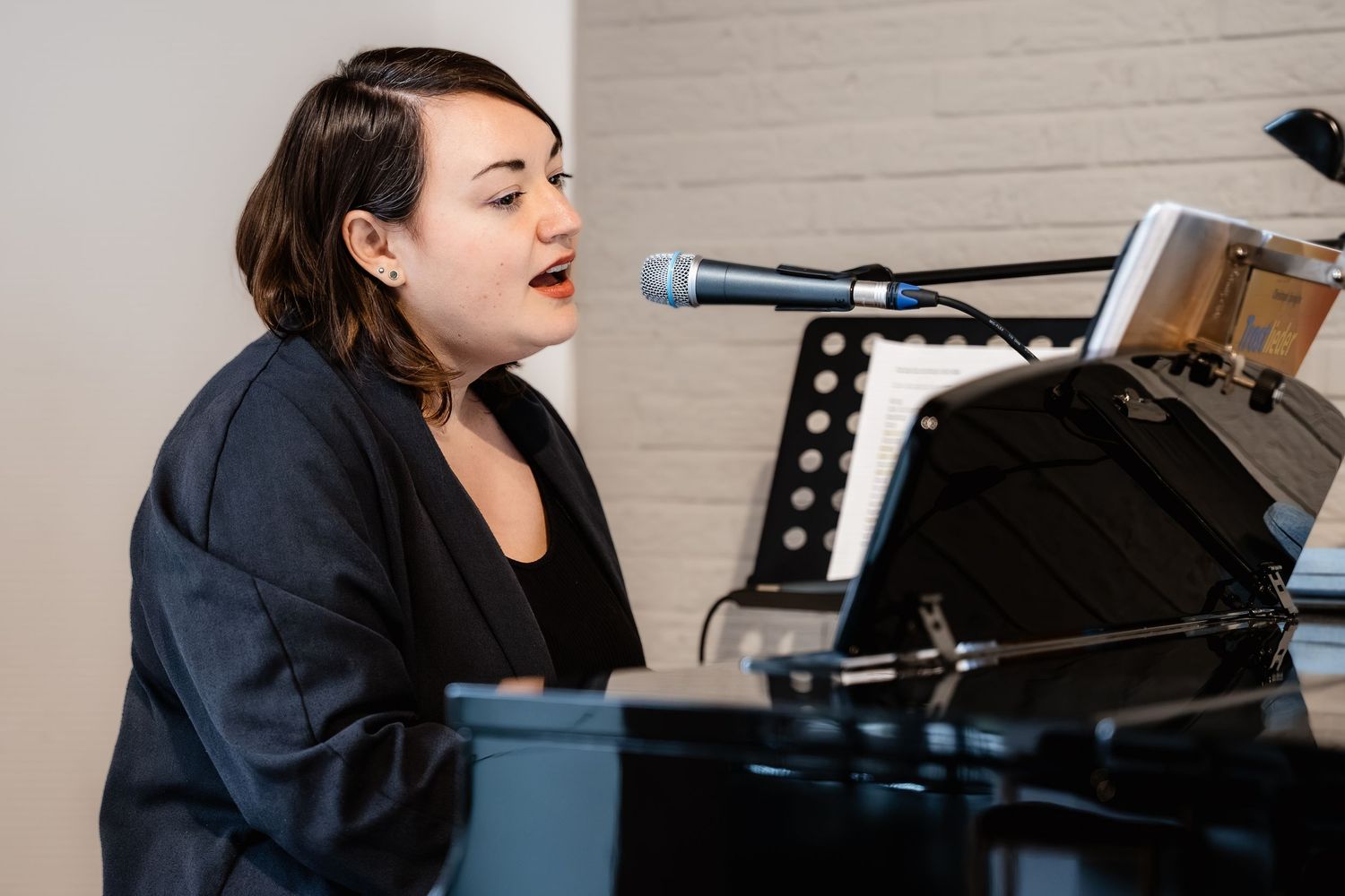 Young woman singing passionately while playing a black grand piano, wearing a dark blazer in a bright studio.