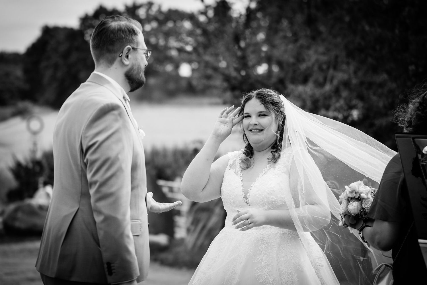 Black and white photo of a bride adjusting her veil while smiling at the groom during an outdoor wedding ceremony.