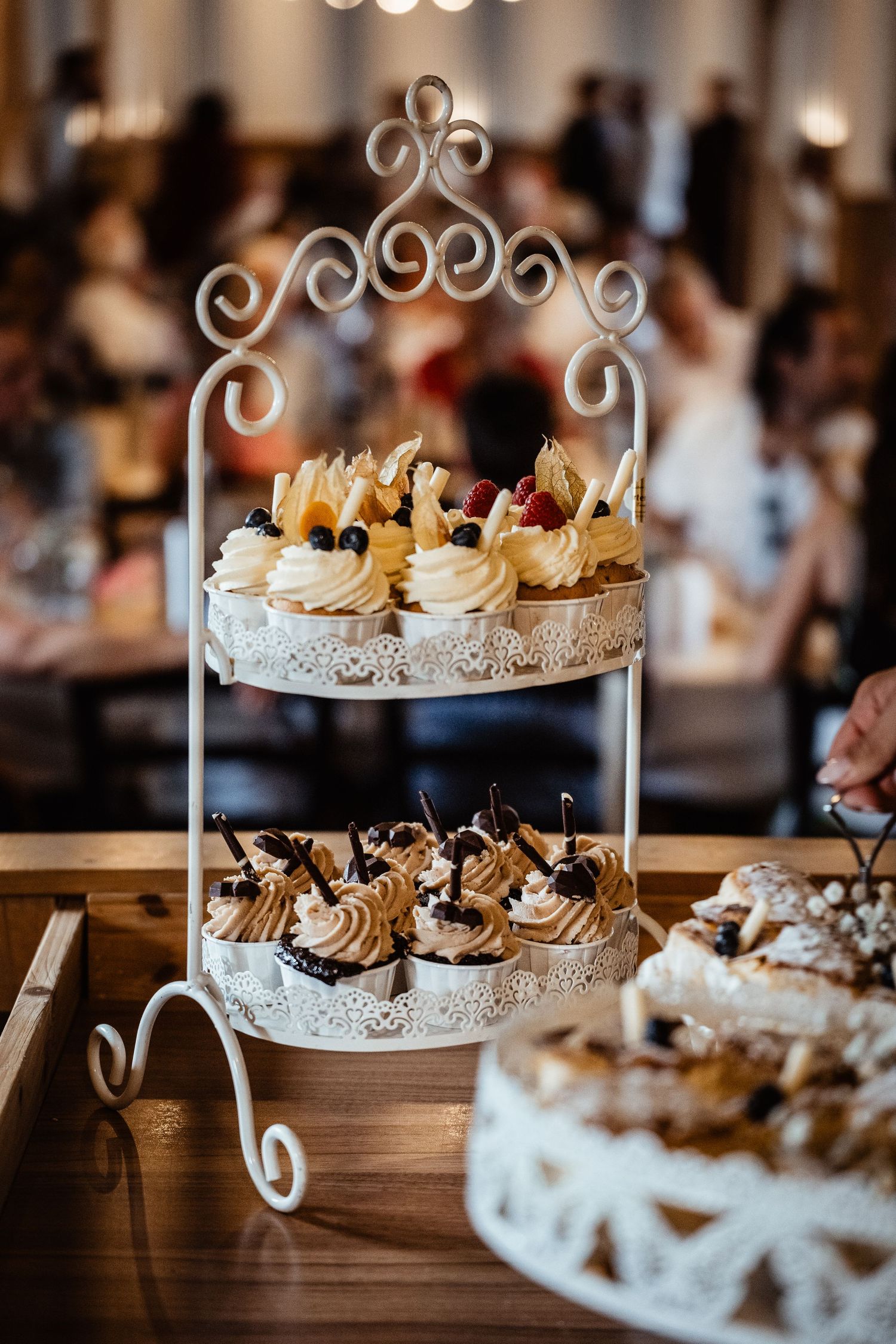 Elegant two-tier white ornate dessert stand displaying assorted cupcakes with cream toppings and fresh fruit at a reception.