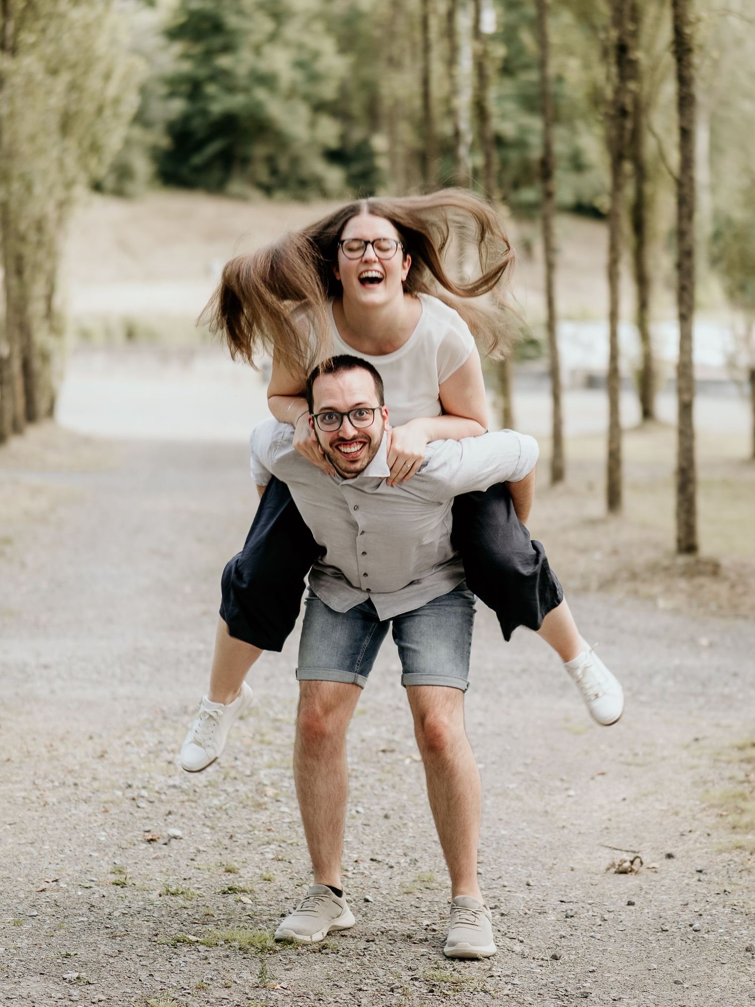 Man giving woman a piggyback ride laughing outdoors on a tree-lined path.