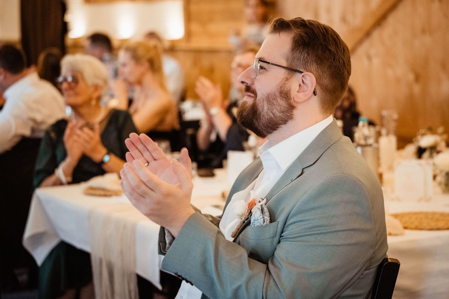Bearded groom in gray suit clapping at wedding reception table with guests in wooden barn venue.