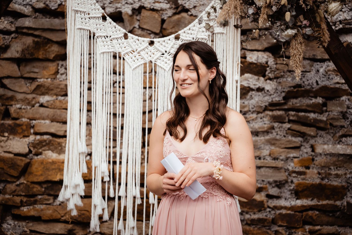 Smiling bridesmaid in pink dress holding notes, standing before rustic stone wall with macrame decor.