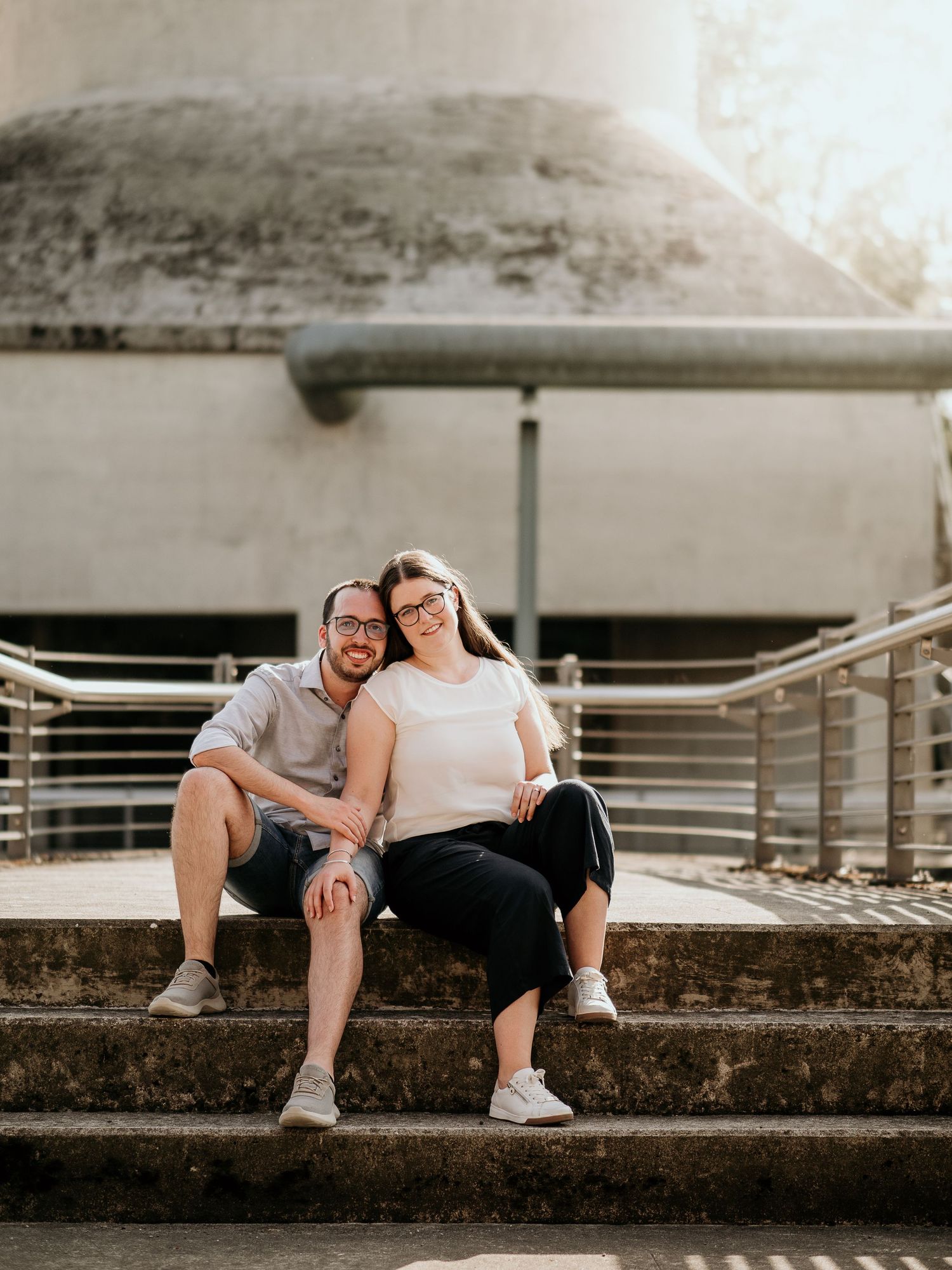A couple sits closely together on concrete steps, smiling warmly, with modern architecture in the background.