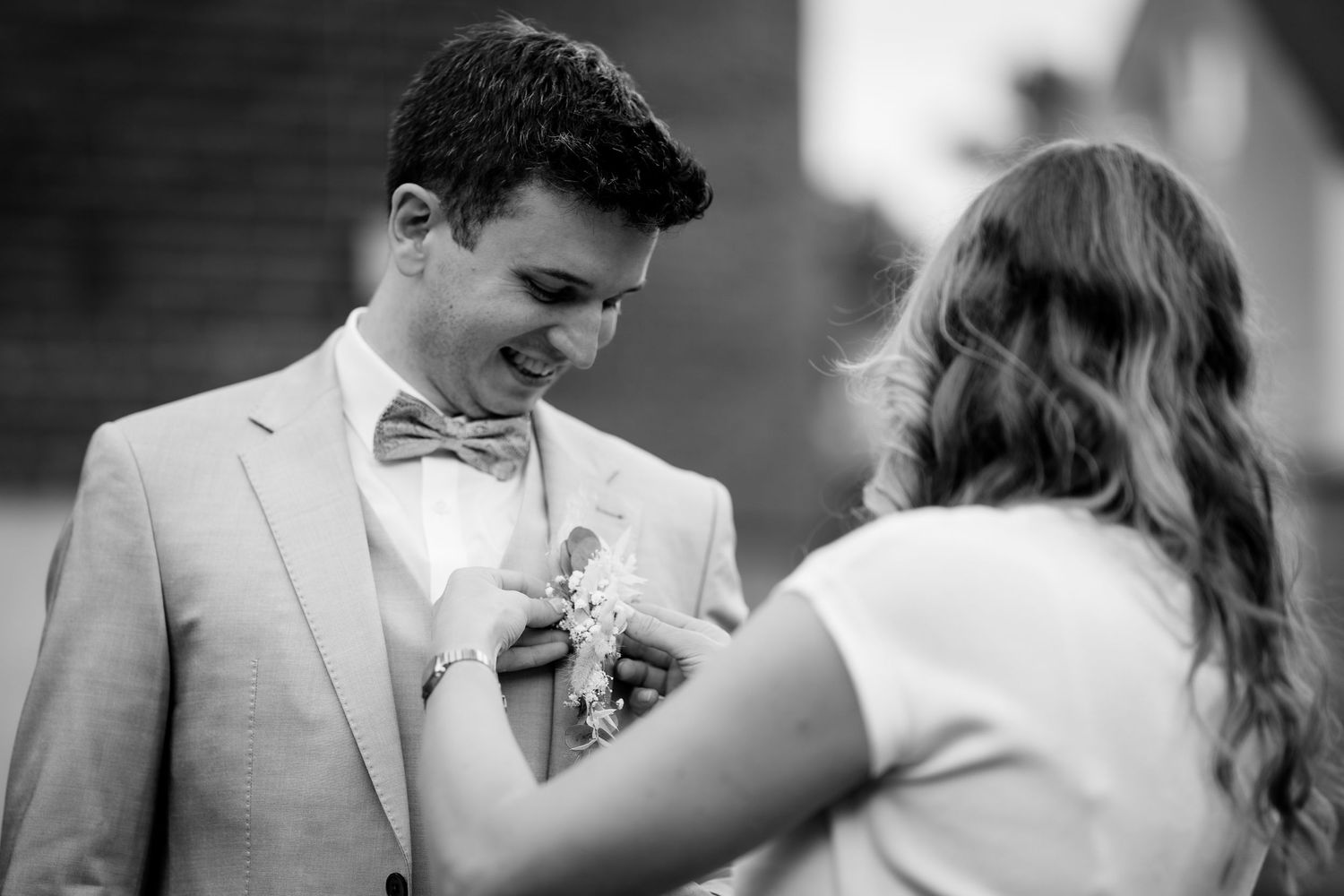 Black and white photo of a woman pinning a boutonniere on a smiling man in a bow tie and suit.