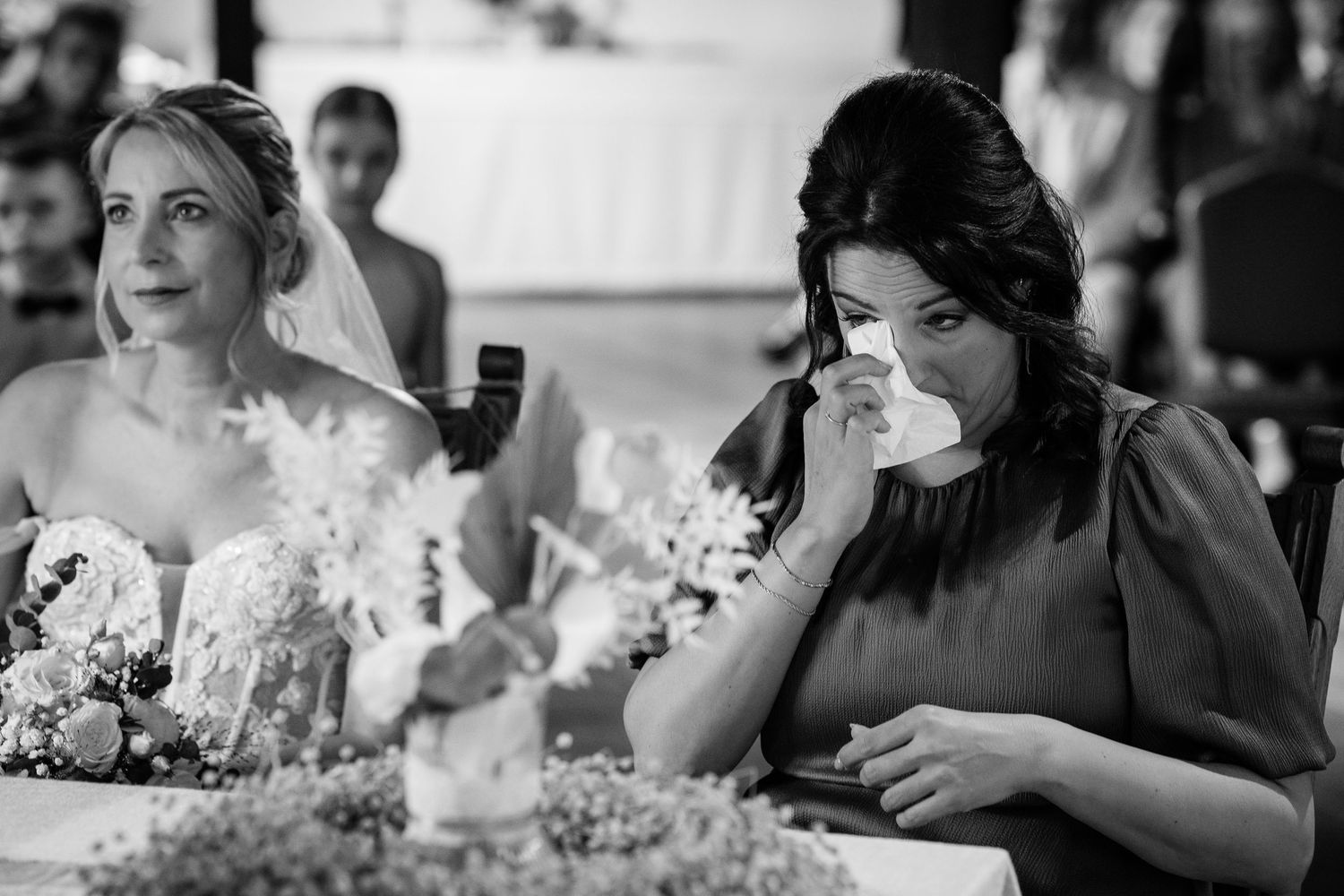 Black and white photo of emotional wedding guest wiping tears while bride sits nearby.