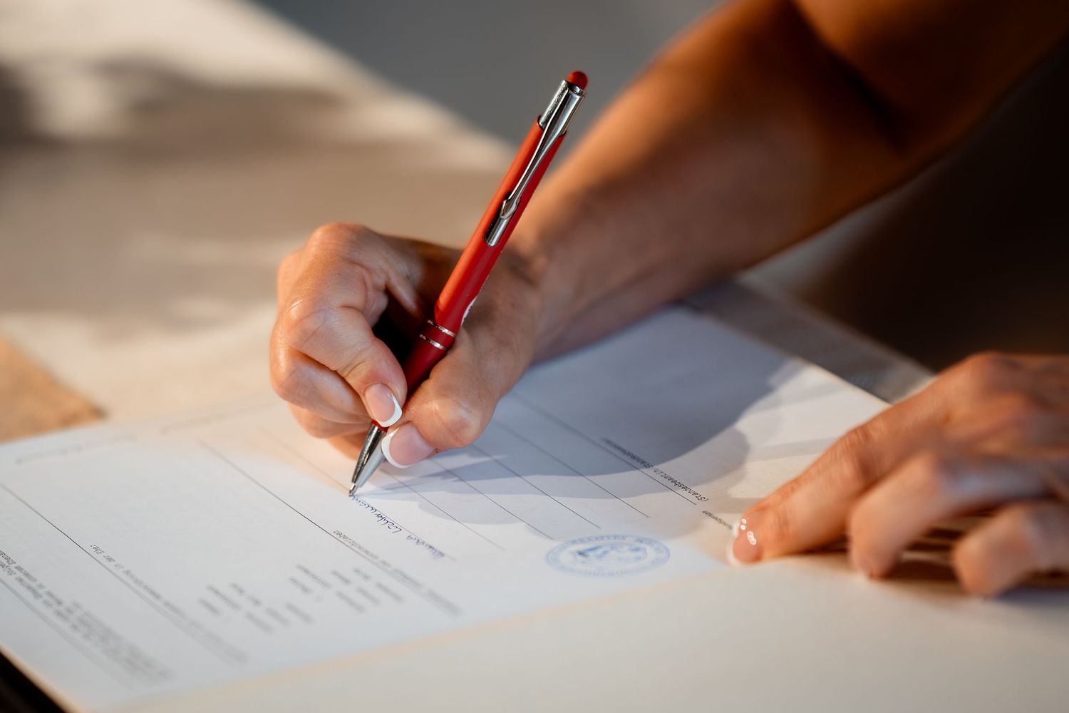Close-up of a hand signing an official document with a red pen on a white paper.