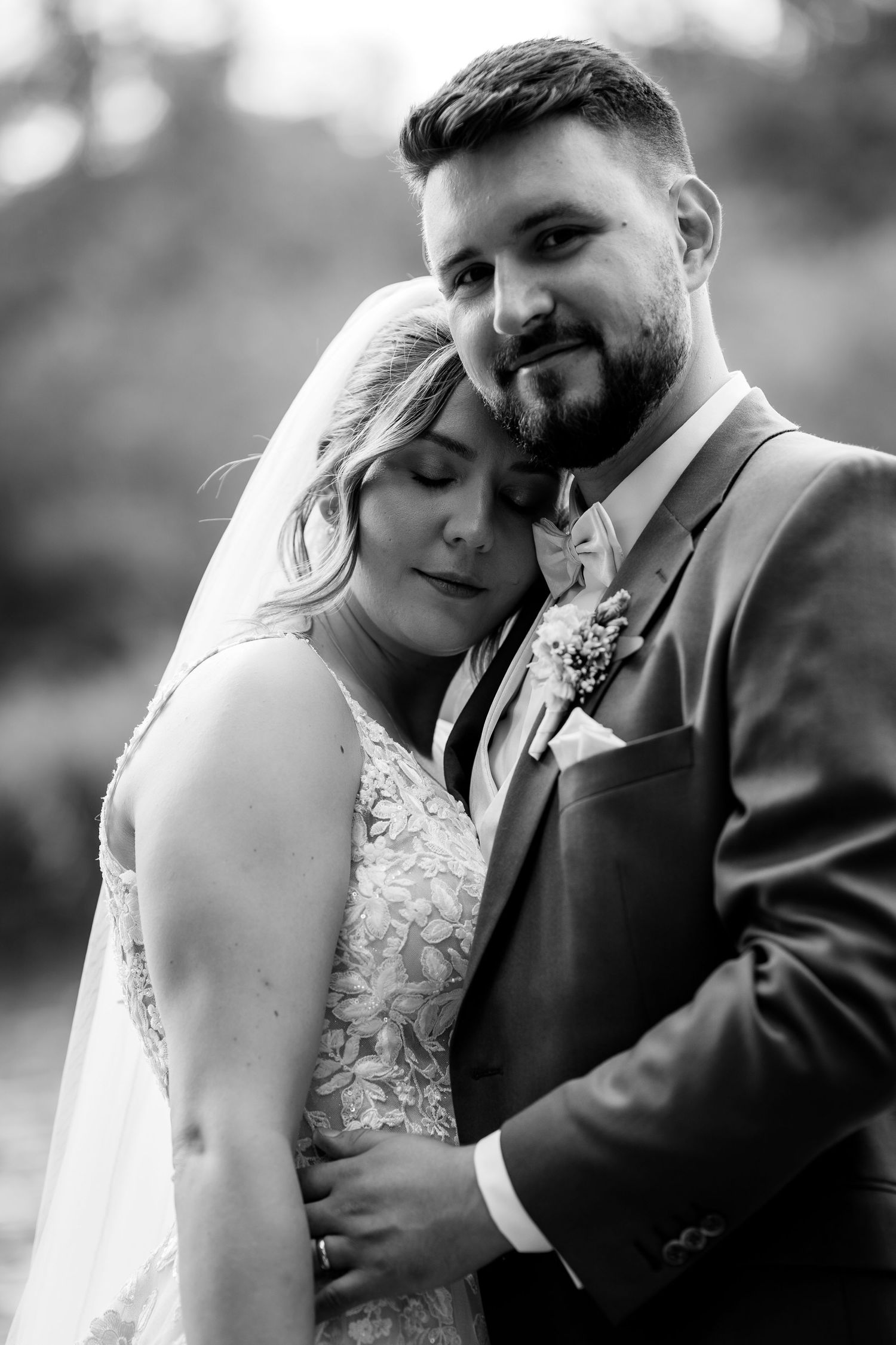 Black and white wedding portrait of bride and groom embracing tenderly, bride in lace gown with veil, groom in suit with boutonniere.