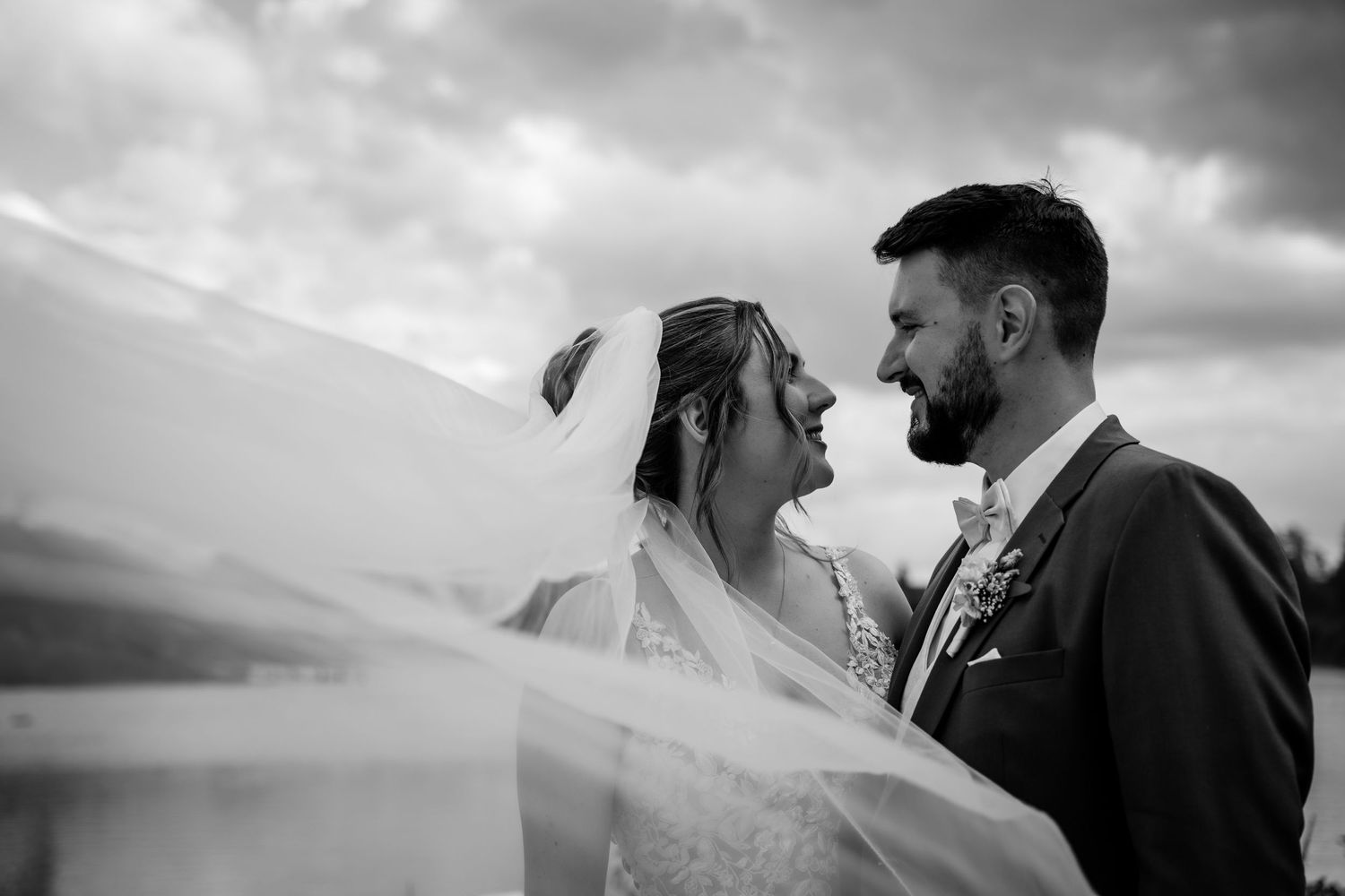 Black and white photo of a bride and groom gazing at each other, veil flowing in the wind against a dramatic cloudy sky.