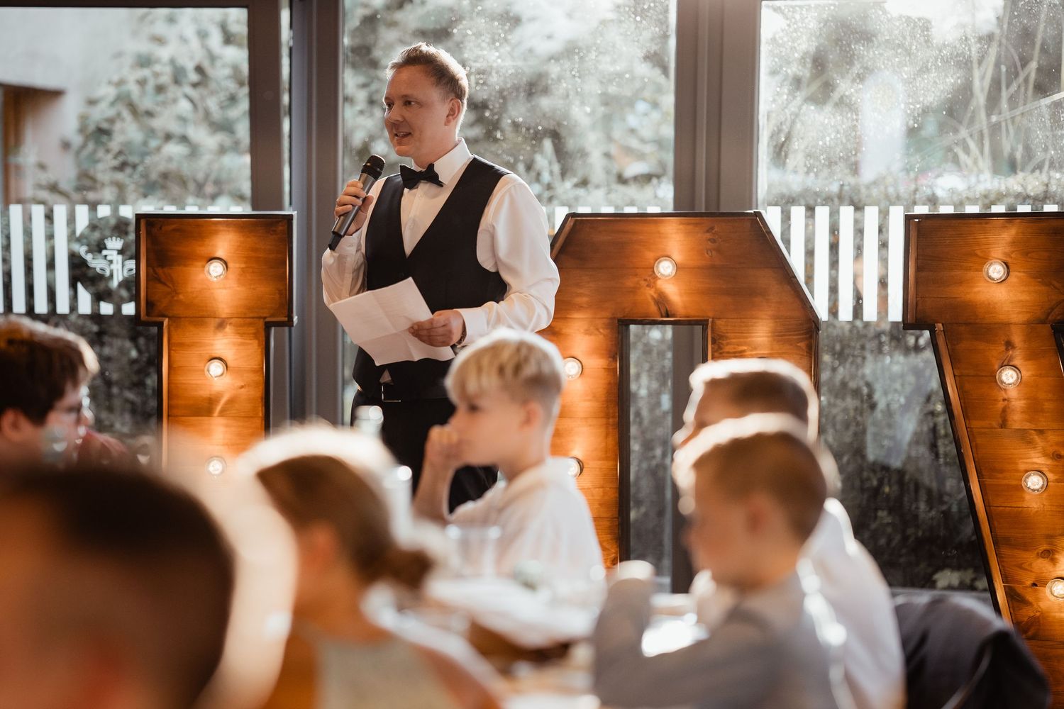 Groom in bow tie delivers wedding speech at reception with illuminated letter decorations.