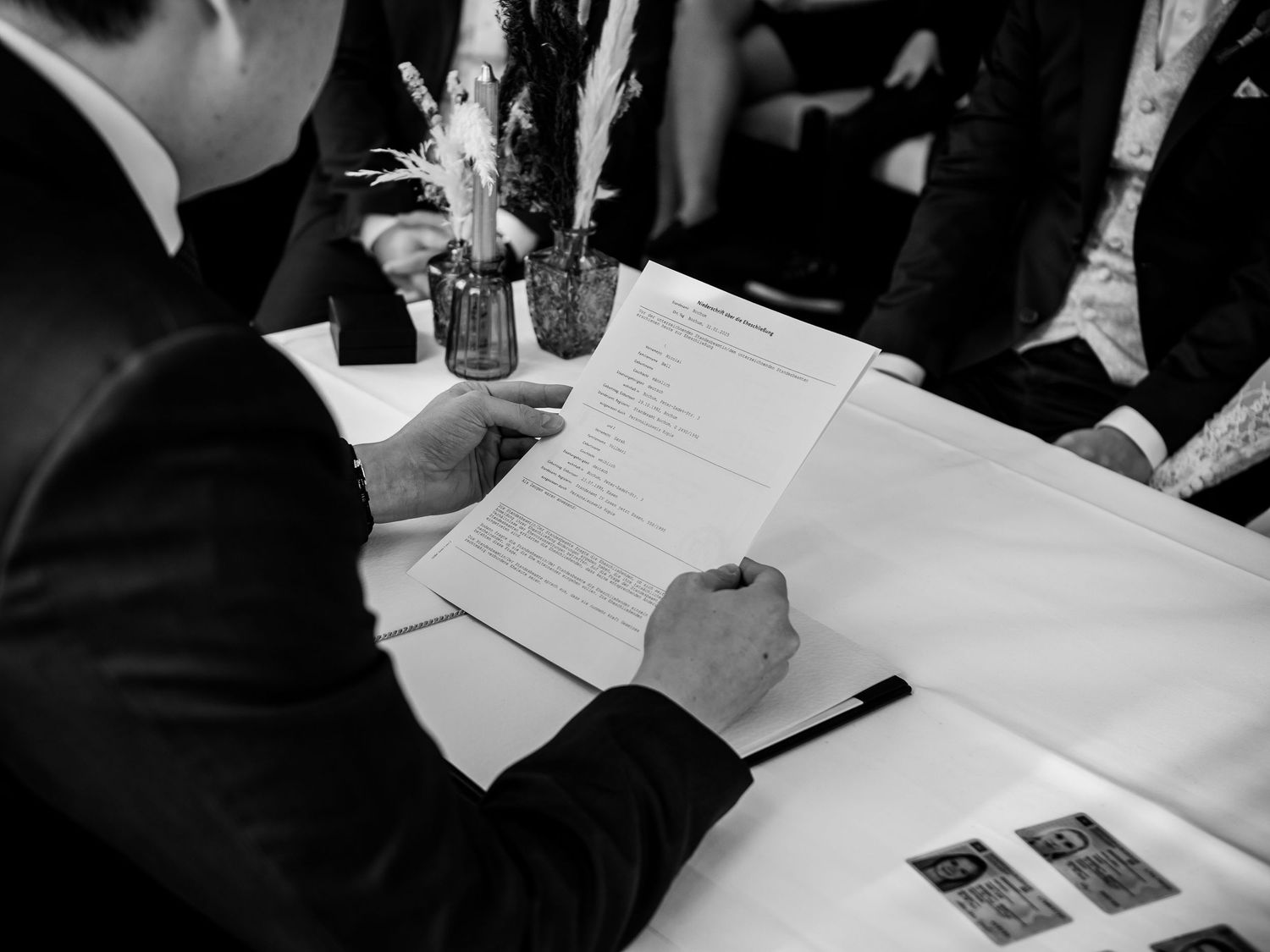 Groom signing the wedding certificate at a table with a floral arrangement in black and white photo.