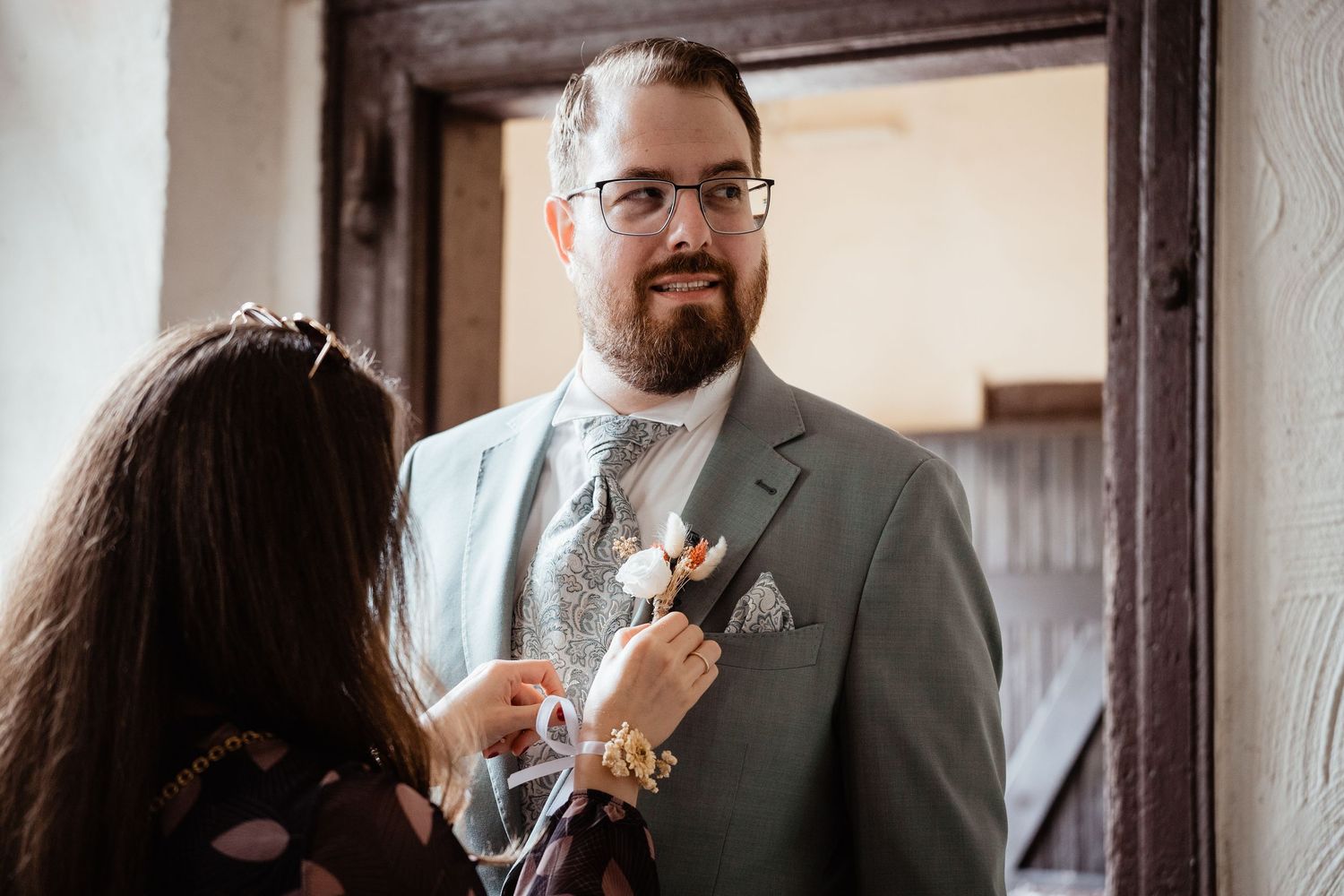 A bearded groom in a sage green suit receives a boutonniere from a woman during wedding preparations.