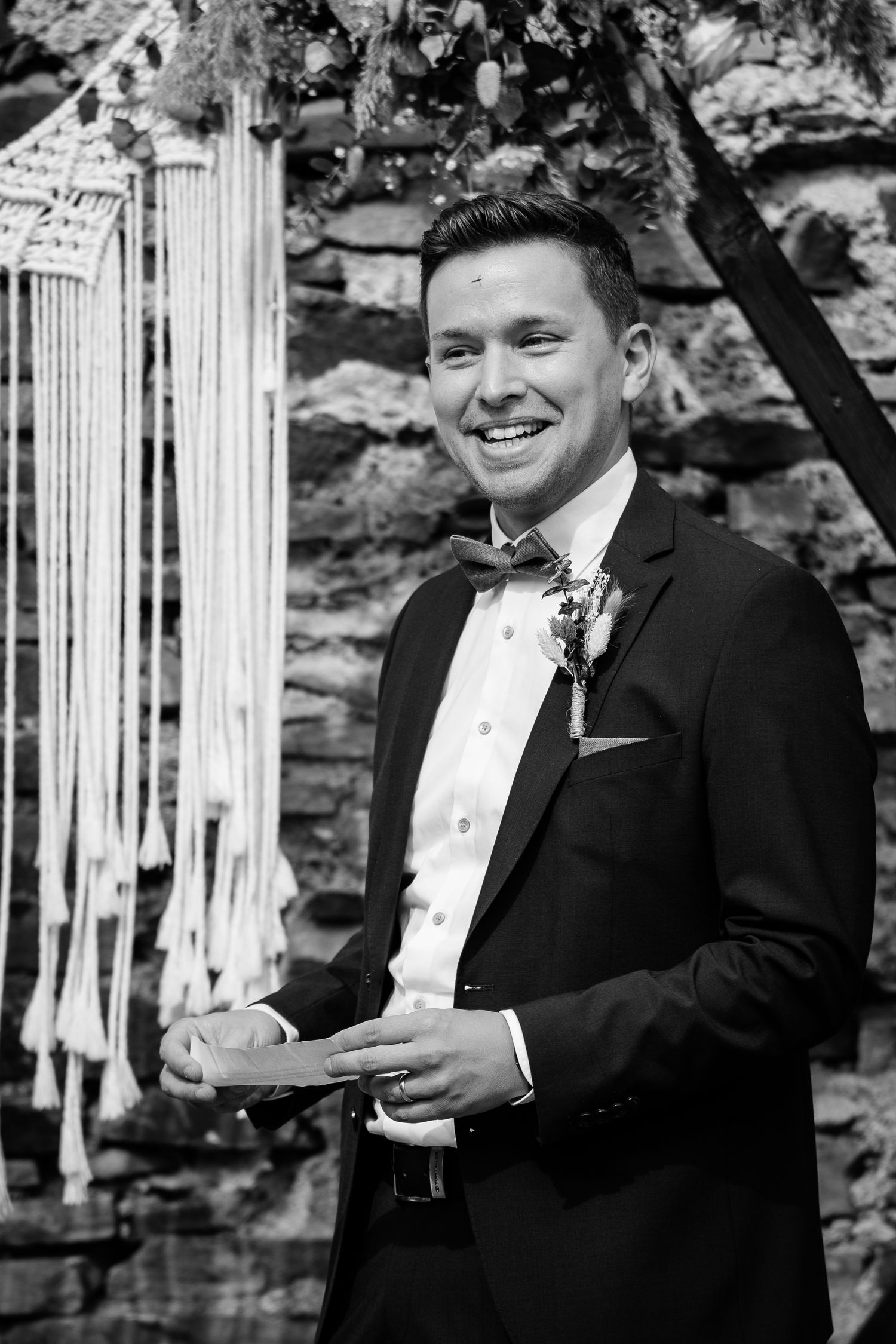Smiling groom in black tuxedo with bow tie holds wedding rings at outdoor ceremony in black and white photo.
