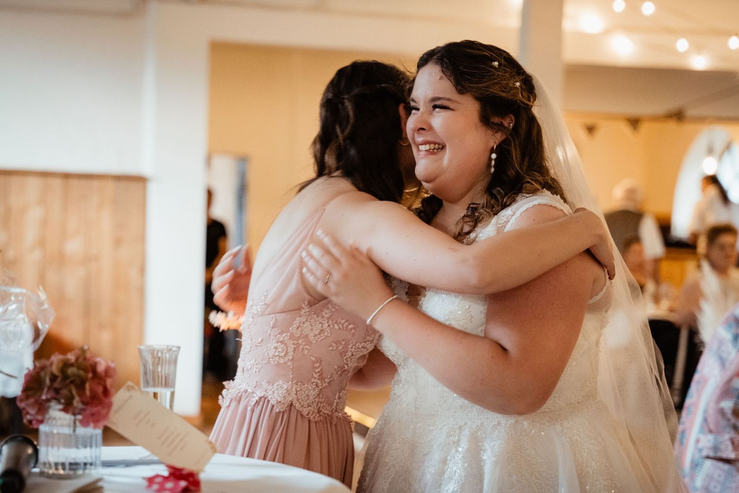 Bride in white lace gown shares a joyful hug with bridesmaid in pink dress at wedding reception.