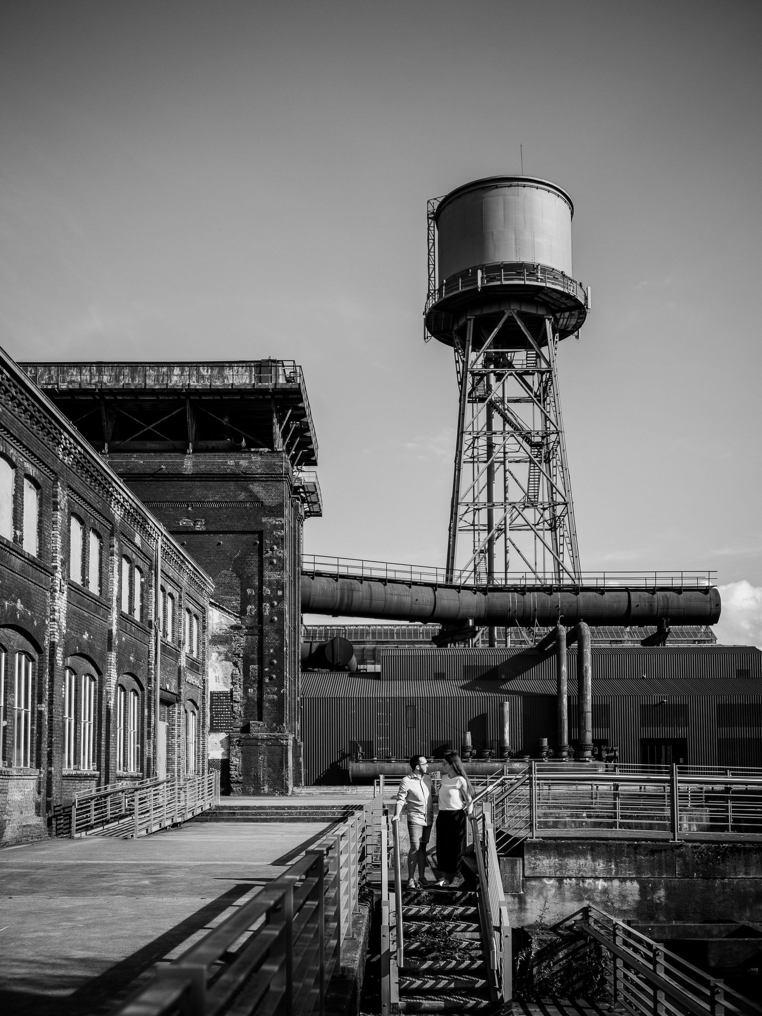 Couple stands on industrial stairs near a vintage water tower at an abandoned factory in black and white.