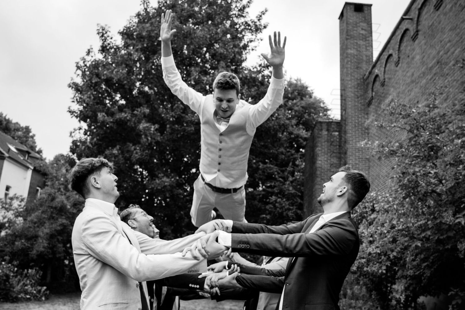 Black and white photo of groomsmen tossing groom into the air outdoors at wedding.