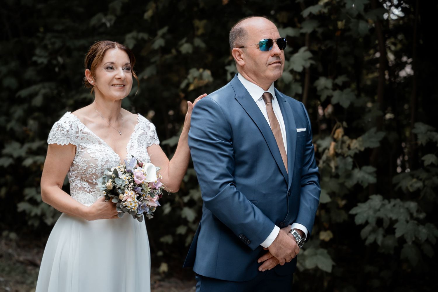 Bride in white lace gown holding bouquet stands beside groom in blue suit with sunglasses outdoors.