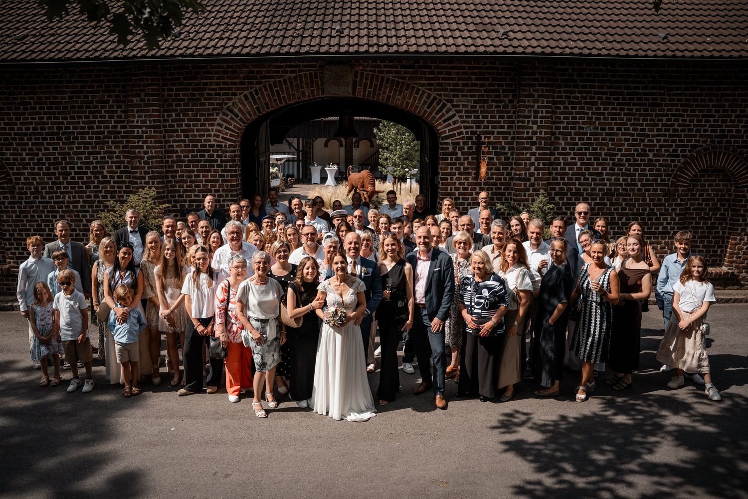 Large wedding group photo with bride and groom surrounded by guests in front of brick archway.
