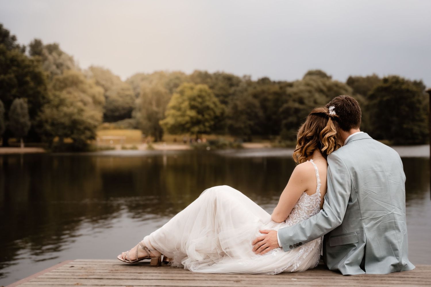 Bride and groom sitting on wooden dock by lake at sunset, surrounded by trees.