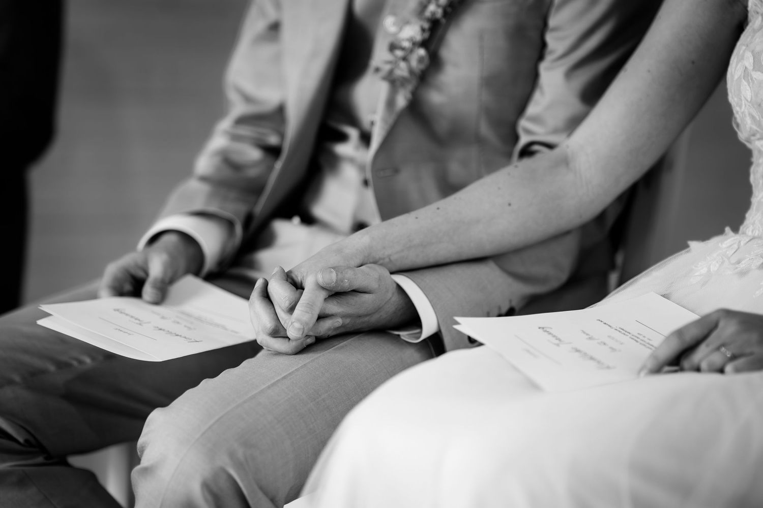 Black and white photo of bride and groom holding hands during wedding ceremony, with programs in their laps.