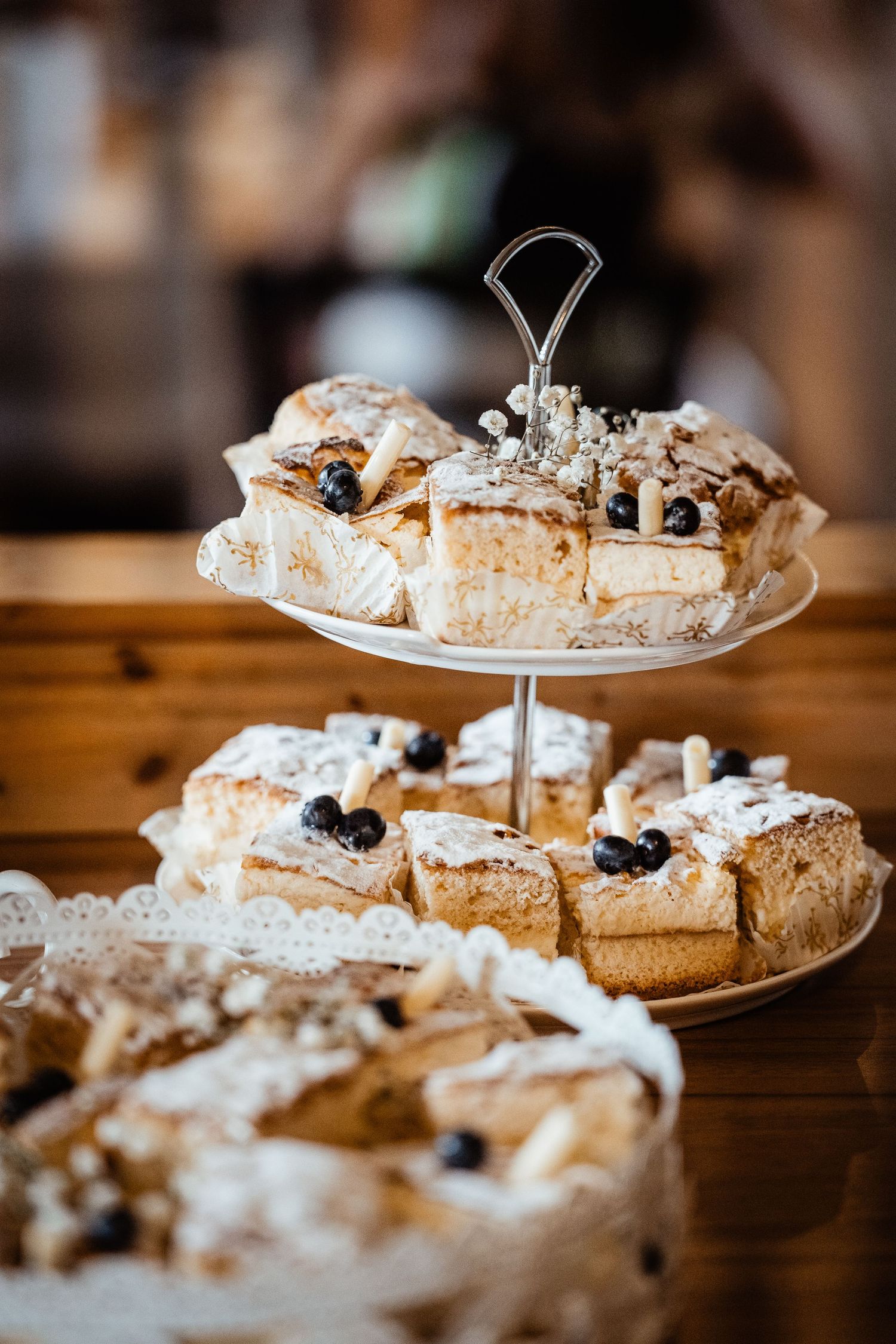 Two-tier white cake stand displaying layered cream pastries topped with blueberries and powdered sugar on a wooden table.