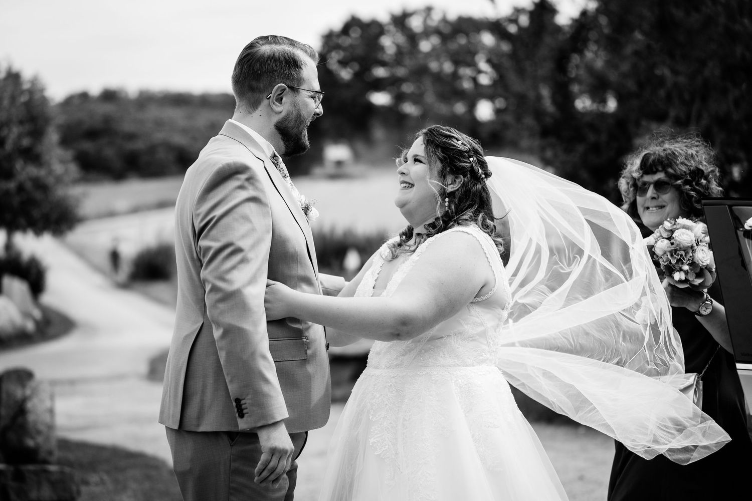 Black and white photo of a bride and groom holding hands during outdoor wedding ceremony, veil blowing in wind.