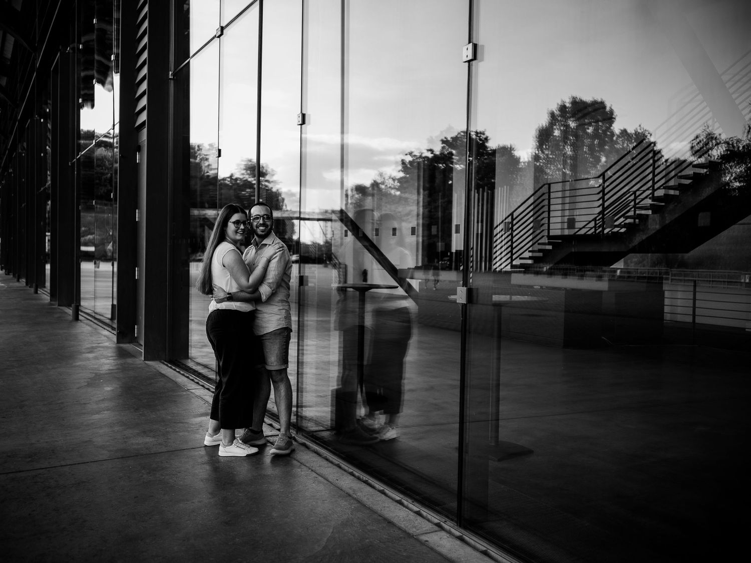 Black and white photo of a couple embracing near a glass building facade at dusk, with their reflection visible.