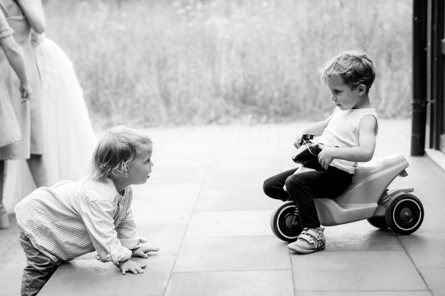 Black and white photo of two young children playing, one on a toy scooter outdoors.