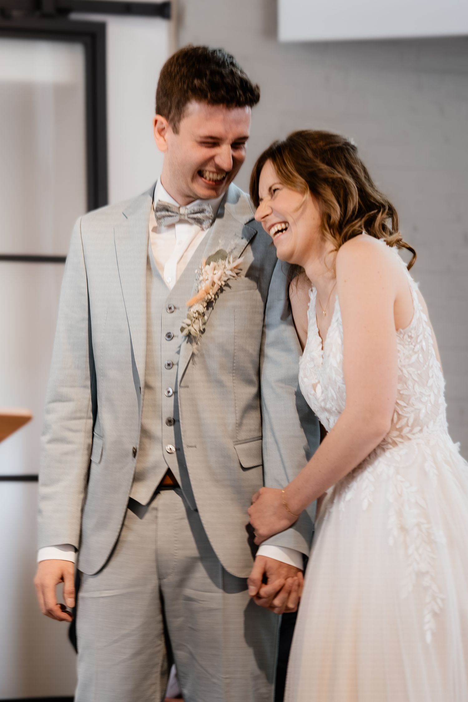 Laughing bride and groom holding hands at wedding ceremony, groom in gray suit with boutonniere.