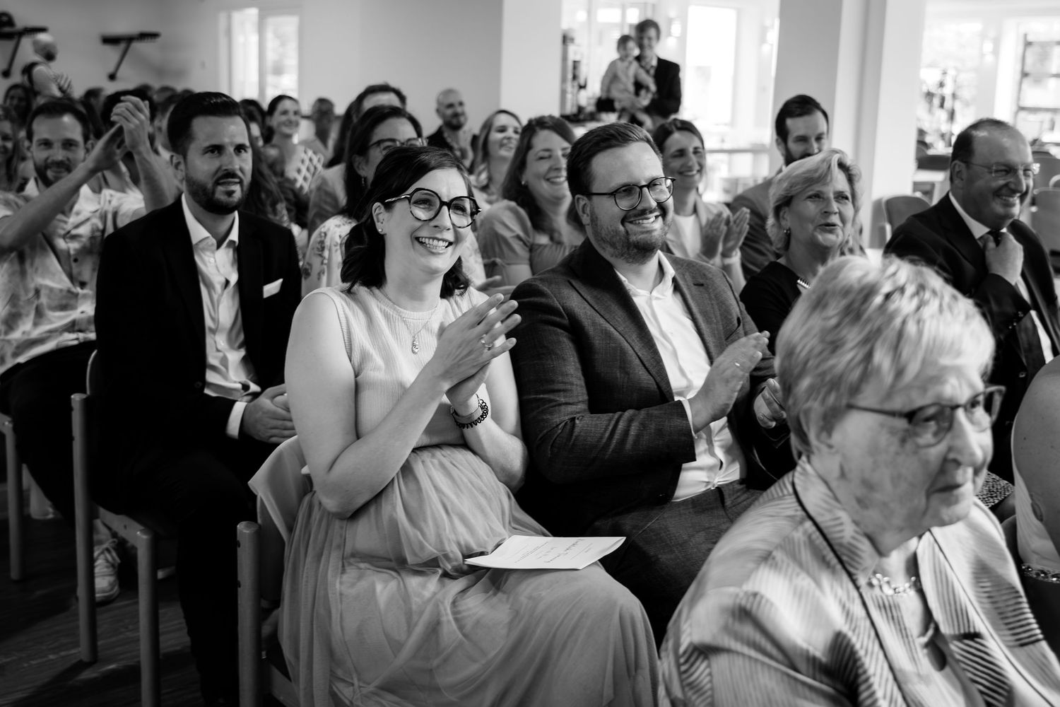 Black and white photo of wedding guests smiling and clapping during a ceremony indoors.