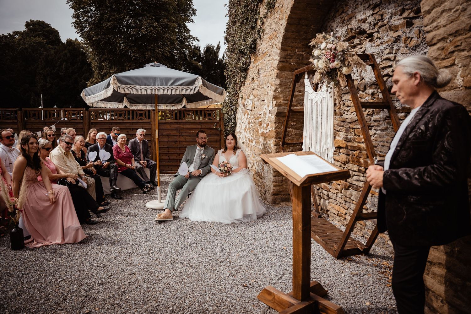 Outdoor wedding ceremony with couple seated before officiant at stone wall venue with guests.