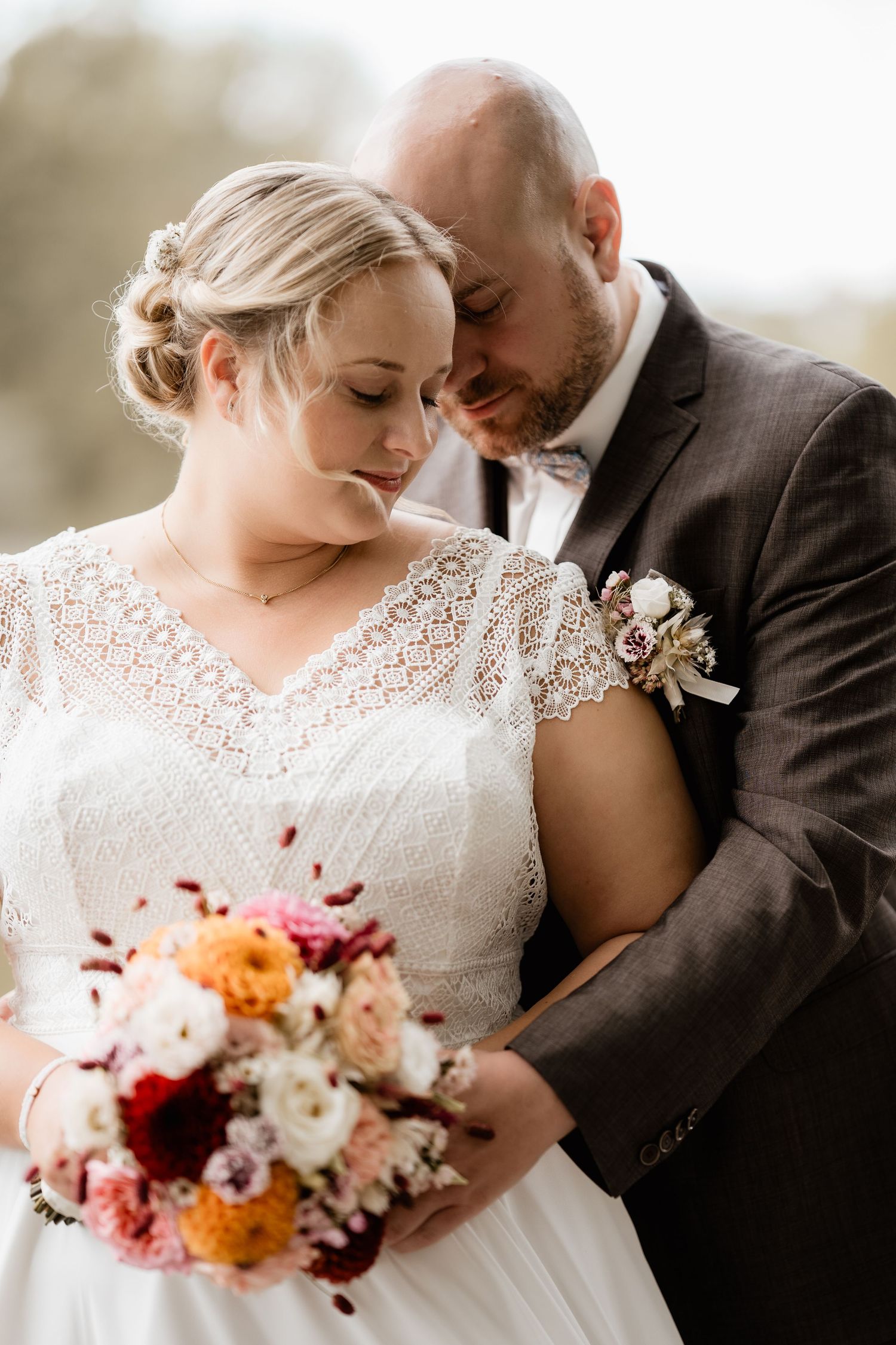 Bride in lace dress holding colorful bouquet embraced by groom in gray suit with boutonniere at wedding.