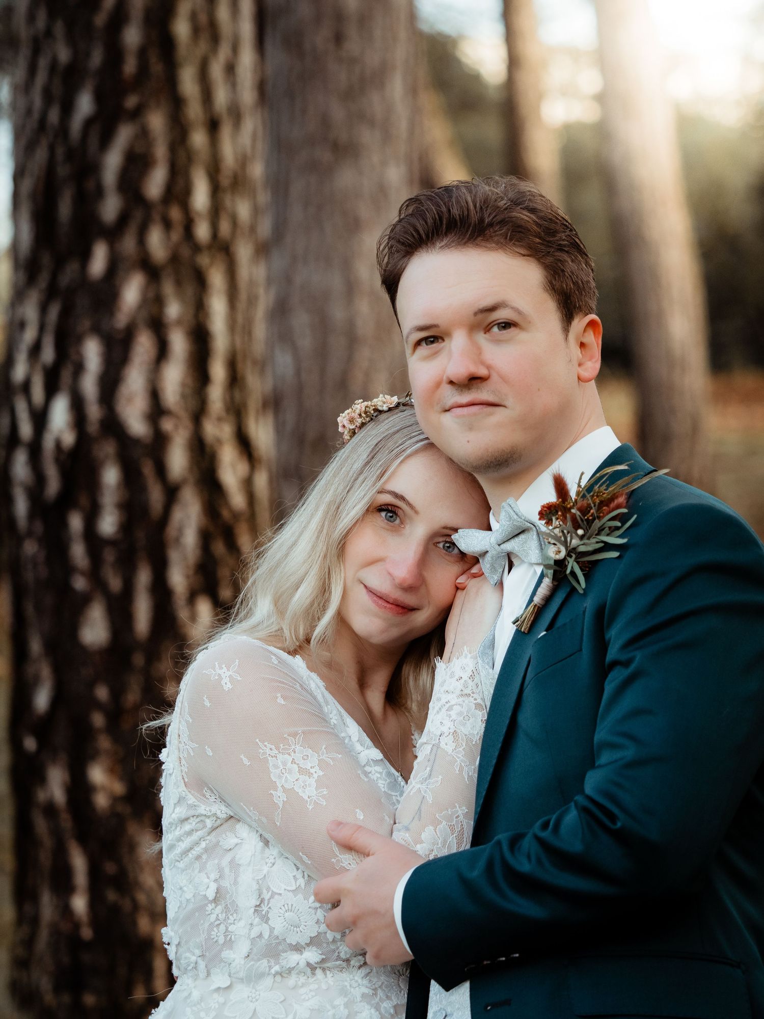 Bride leaning on groom's shoulder in a forest, both wearing wedding attire with warm sunlight behind them.