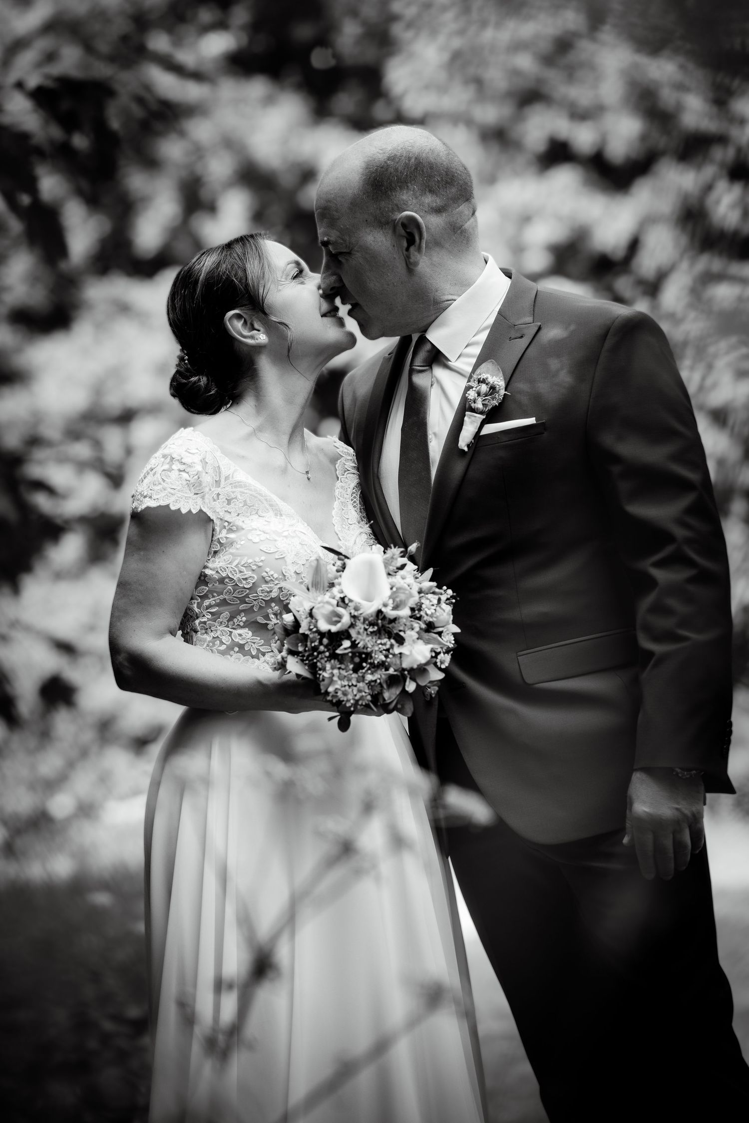 Bride and groom share intimate moment holding bouquet in black and white wedding portrait outdoors.