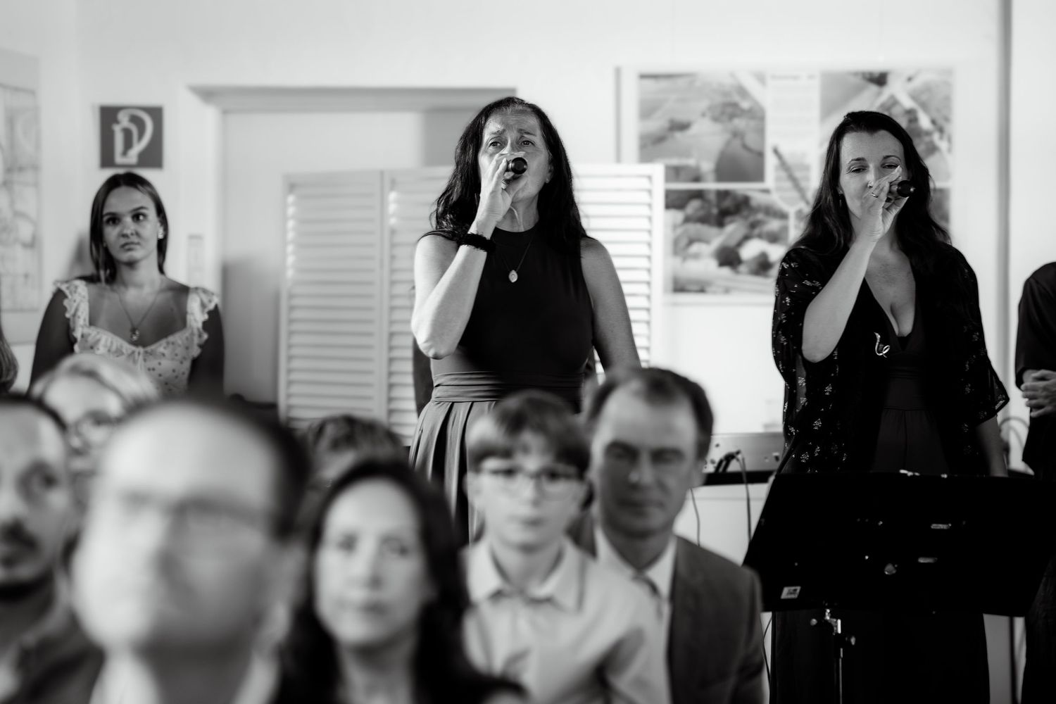 Three women singing and performing at a wedding ceremony while guests watch in a bright white venue.