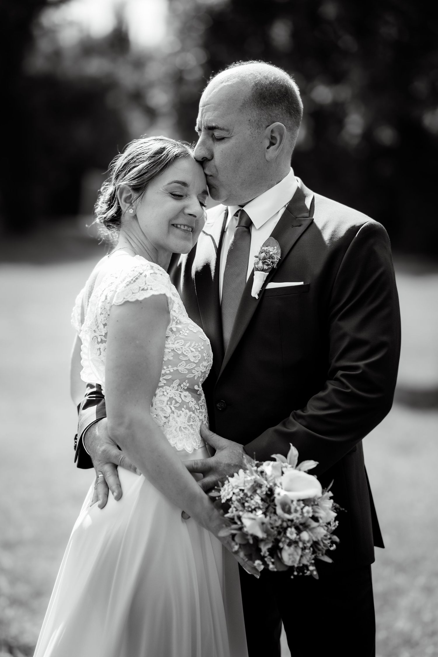 Black and white wedding photo of groom kissing bride on forehead while she smiles holding a bouquet outdoors.
