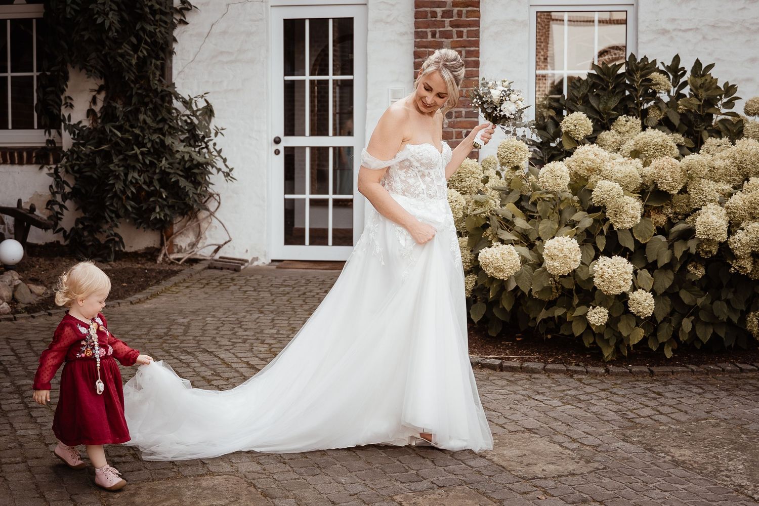 Bride in white gown with toddler in red dress near hydrangeas outside venue.