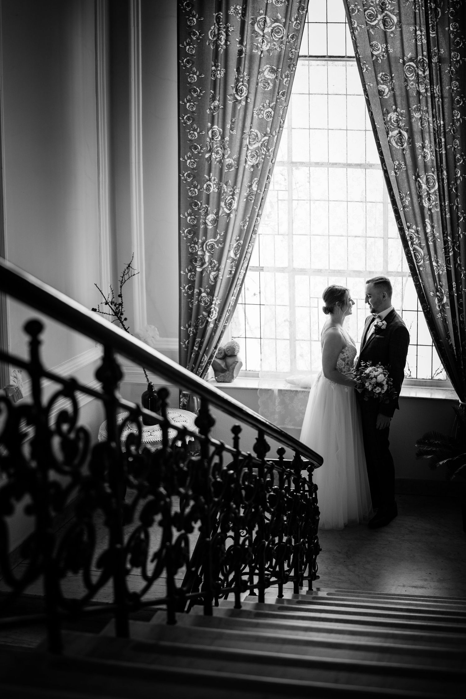 Black and white wedding photo of bride and groom standing by grand staircase window with elegant curtains.