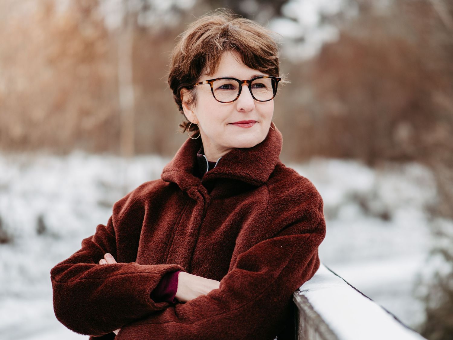 A child in a burgundy winter coat poses thoughtfully in a snowy winter landscape with blurred trees in the background.