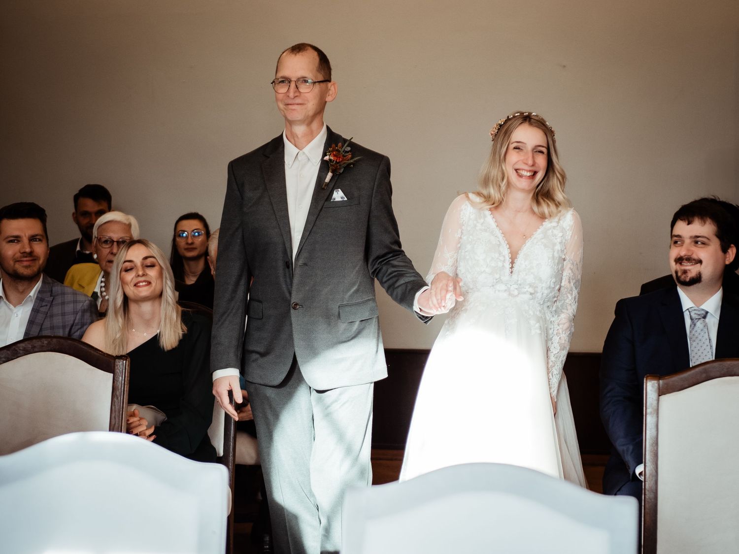 Bride in white gown walks down aisle with escort at wedding ceremony, guests smiling in background.