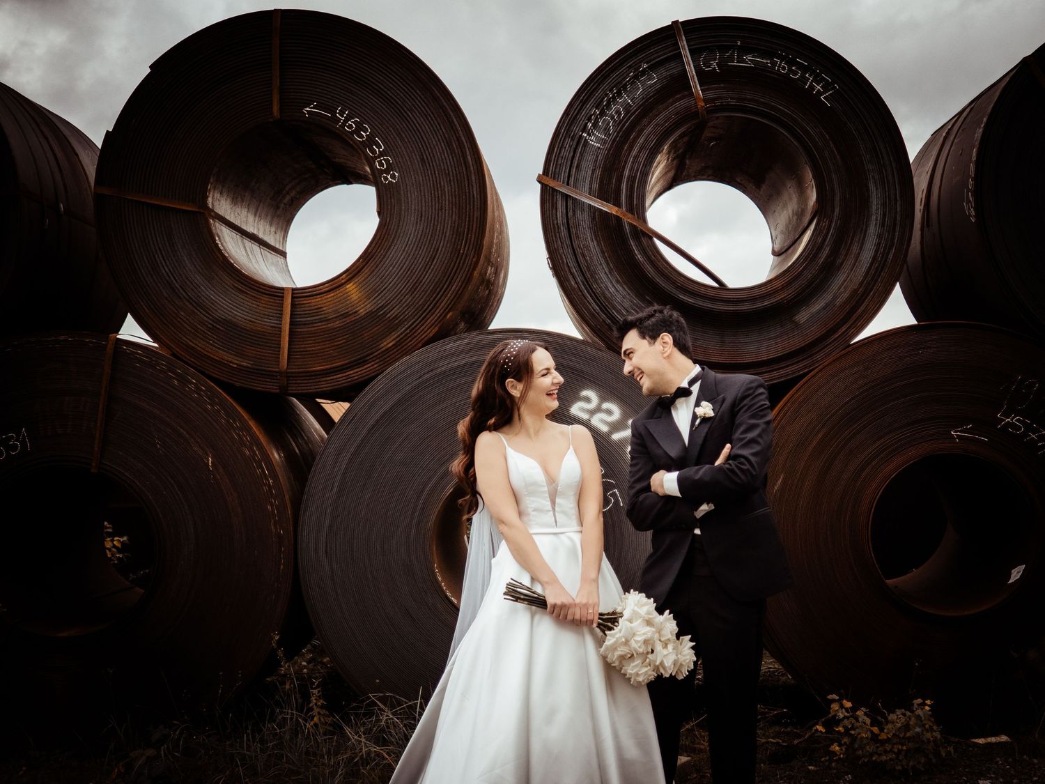 Bride and groom pose romantically before large industrial steel coils in a unique wedding photo.