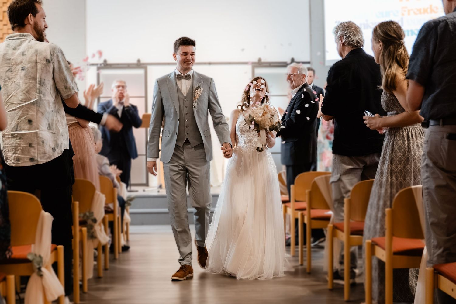 A happy bride and groom walk hand in hand down the aisle as guests celebrate around them.
