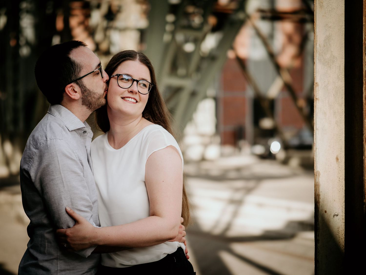Couple sharing a romantic embrace outdoors, woman in white top with glasses smiling warmly near industrial structure.