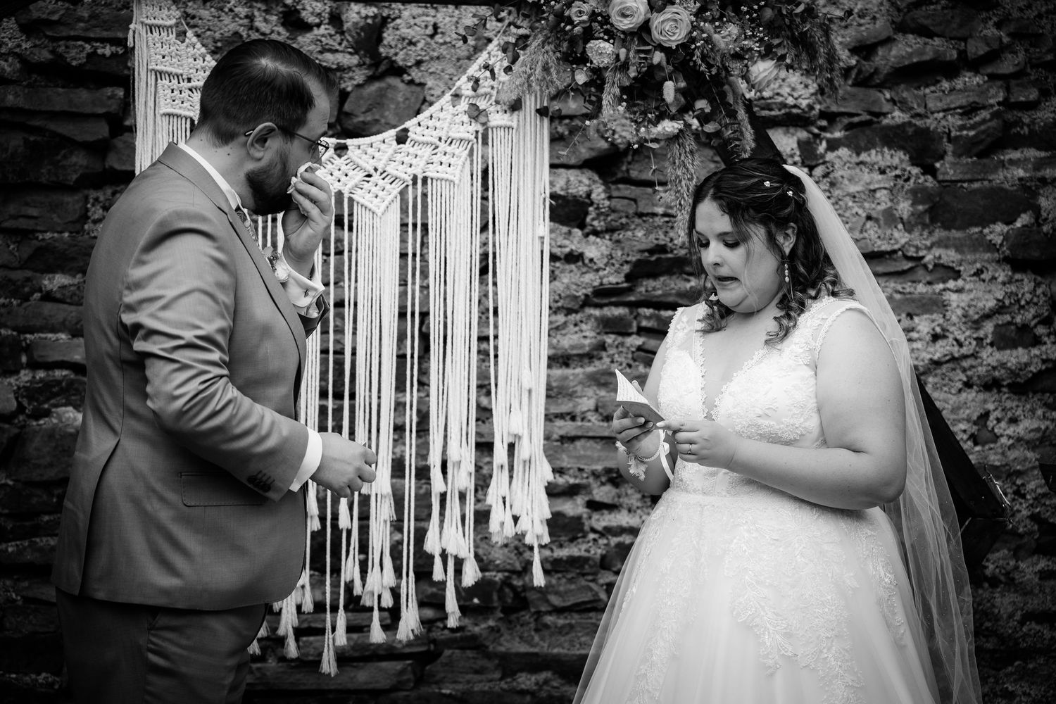 Black and white photo of bride and groom exchanging vows before a macrame backdrop in a stone chapel.
