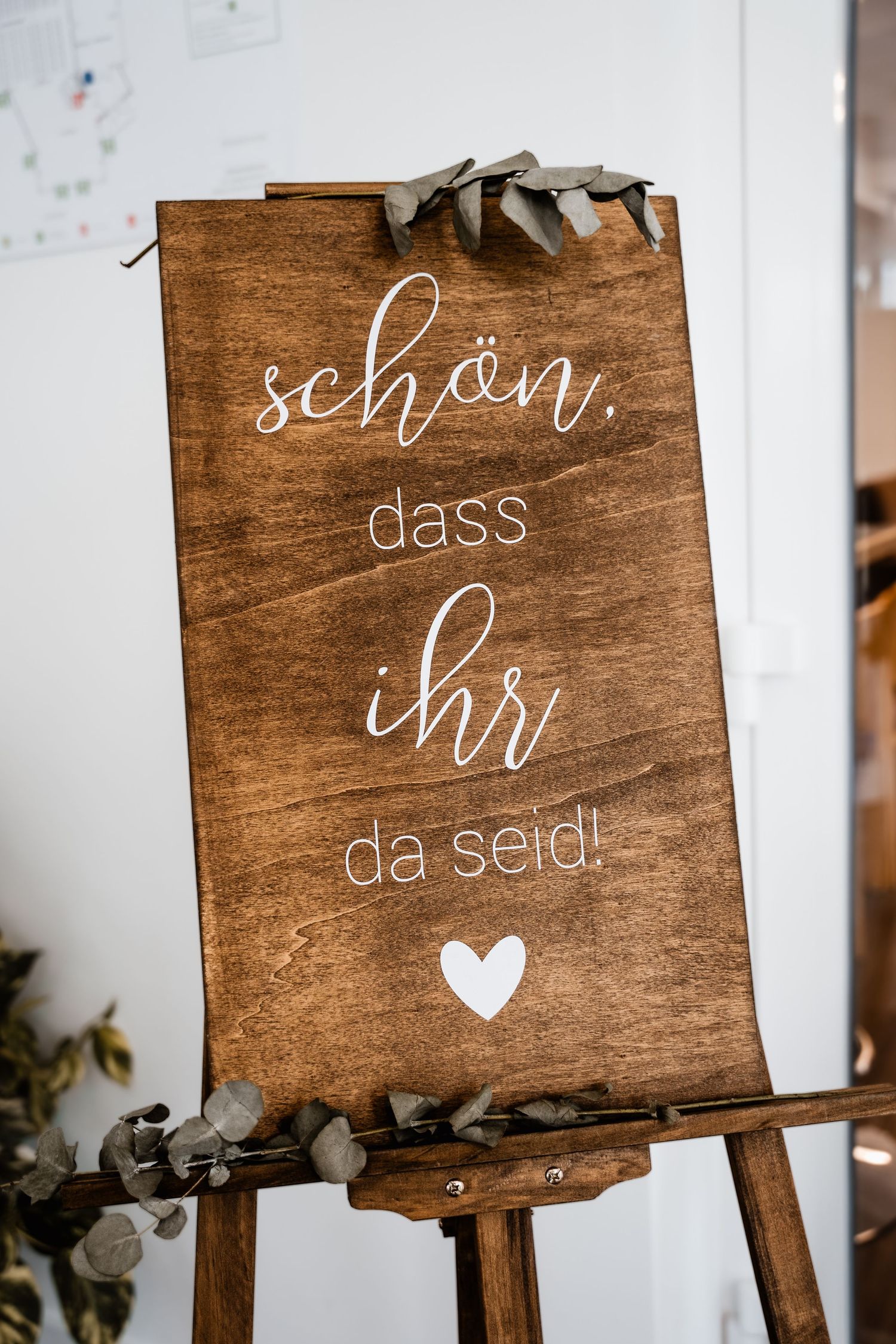 A rustic wooden wedding welcome sign reading schon dass ihr da seid on an easel with eucalyptus decor.