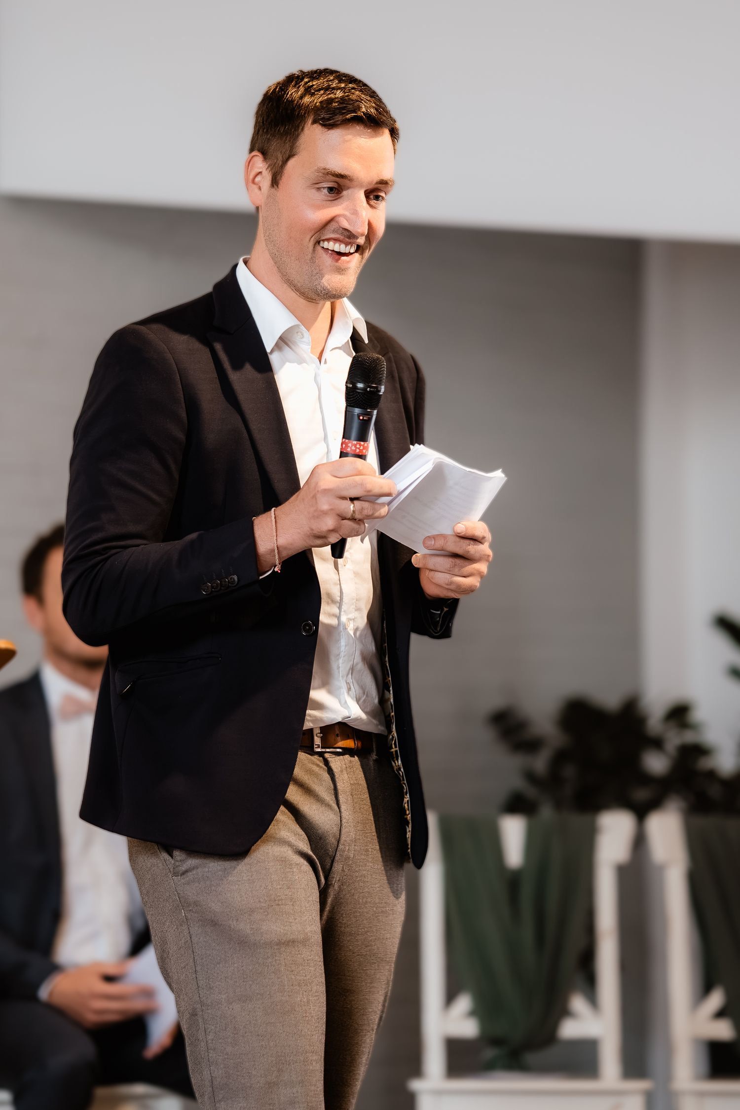 Smiling man in black blazer holding microphone and papers while speaking at a wedding ceremony event.