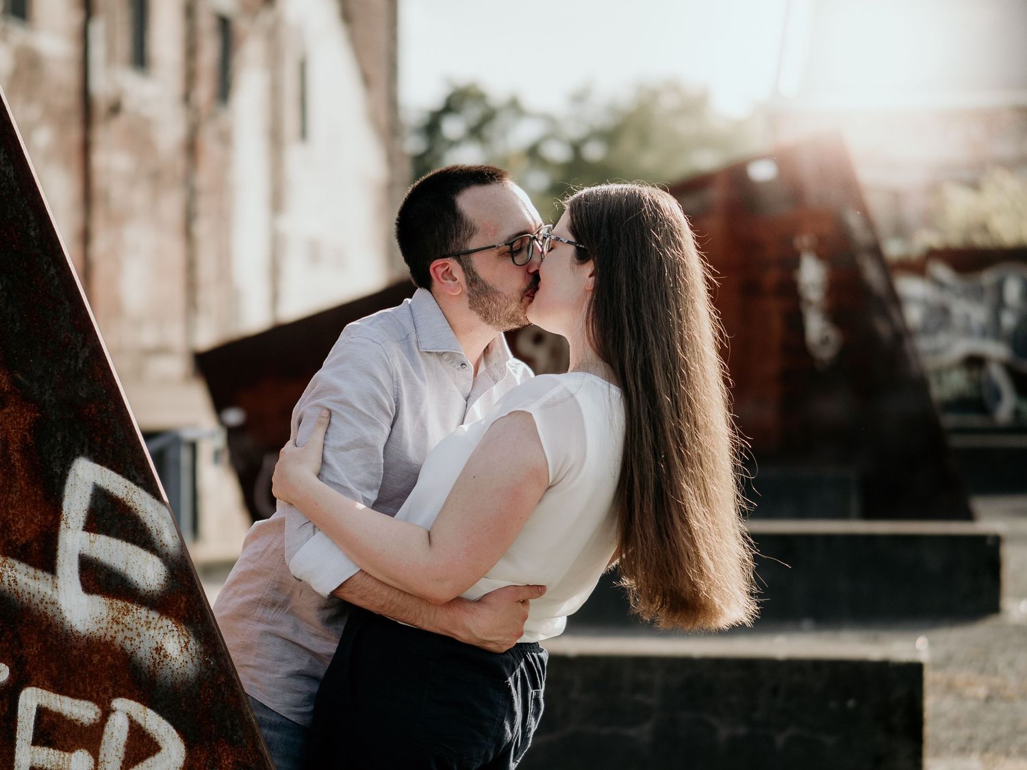 Couple sharing a passionate kiss outdoors with warm sunlight and rustic graffiti walls in the background.