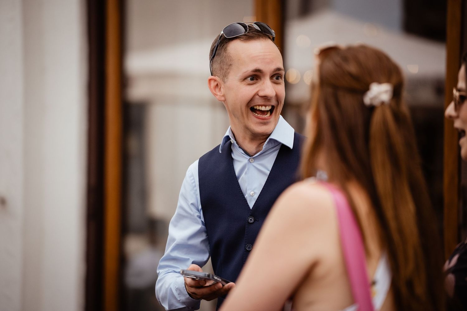 A laughing man in a blue vest and shirt holds a phone while chatting with a woman at a wedding reception.
